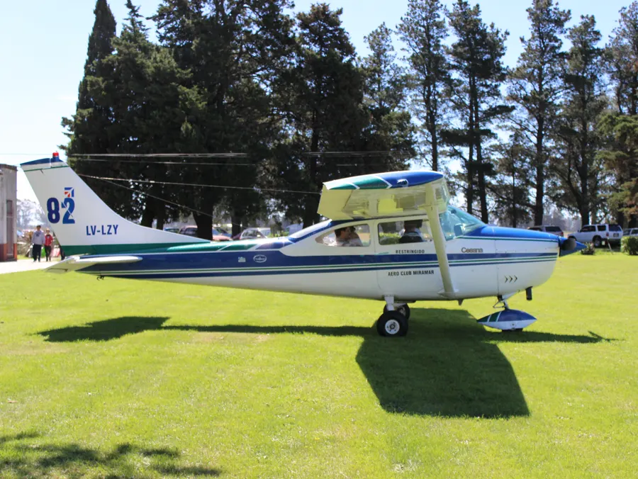La Cessna 182 LV-LZY del Aero Club Miramar estacionada sobre el pasto, blanca con franjas azul y verde y un 82 en la cola
