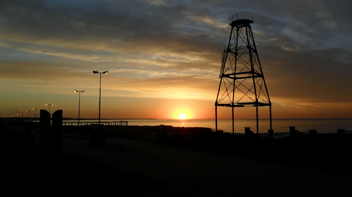 La torre oxidada recortada contra el amanecer sobre el mar, con el muelle y las luces de la costanera a la izquierda