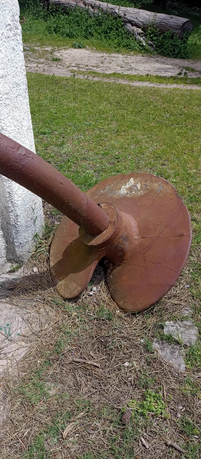 Un caño de hierro oxidado que termina en un disco helicoidal, apoyado en el pasto junto a un pilar blanco