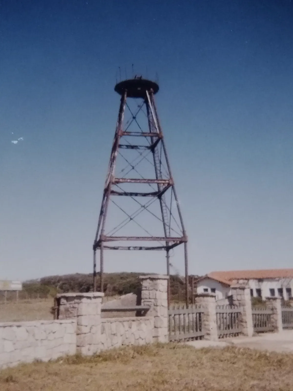 La torre de hierro oxidada, sin linterna ni tablas, detrás de los pilares de piedra y el cerco de la entrada del vivero