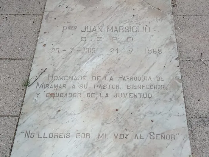 Losa de mármol en la vereda: Pbro. Juan Marsiglio, Q.E.P.D., 23-7-1915, 24-7-1968. Homenaje de la Parroquia de Miramar a su pastor, bienhechor y educador de la juventud. No lloréis por mí, voy al Señor