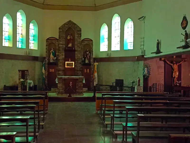 La nave vista desde el fondo: dos filas de bancos de madera, el altar y el retablo de ladrillo al frente, ventanas en punta arriba y, a la derecha, la nave lateral con un Cristo crucificado y la Virgen de Luján