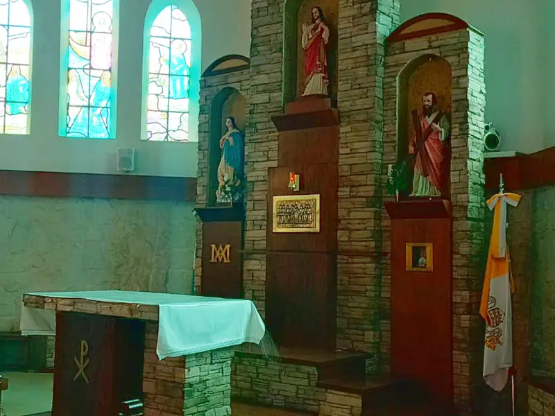 El presbiterio revestido en piedra y ladrillo, con tres hornacinas: el Sagrado Corazón al centro, la Virgen a la izquierda y San Andrés con su cruz a la derecha; adelante, el altar con el monograma de Cristo