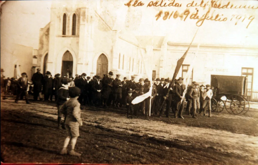 Foto sepia de una multitud de hombres con sombrero y banderas saliendo de la iglesia, con la anotación manuscrita: salida del Tedeum, 9 de julio de 1919