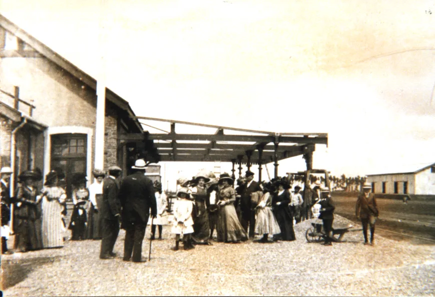 Foto antigua del andén de la estación Miramar: un edificio de ladrillo con la marquesina de madera, un grupo de señoras con sombreros grandes y vestidos largos, hombres de traje y chicos, una carretilla y, al fondo, un galpón largo junto a las vías