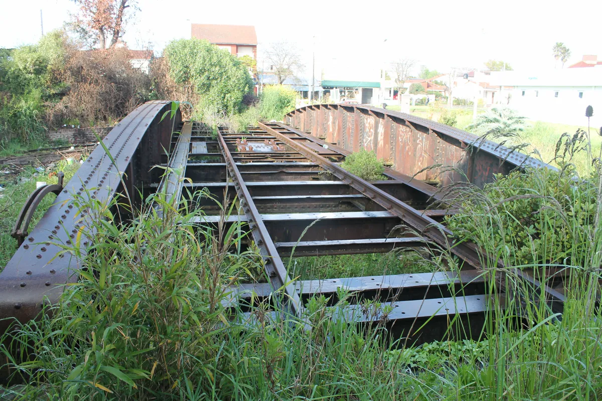 El puente de la mesa giratoria hoy: dos vigas de acero remachadas, oxidadas y con grafitis, más altas en el medio, con la vía sobre el tablero y pasto crecido alrededor