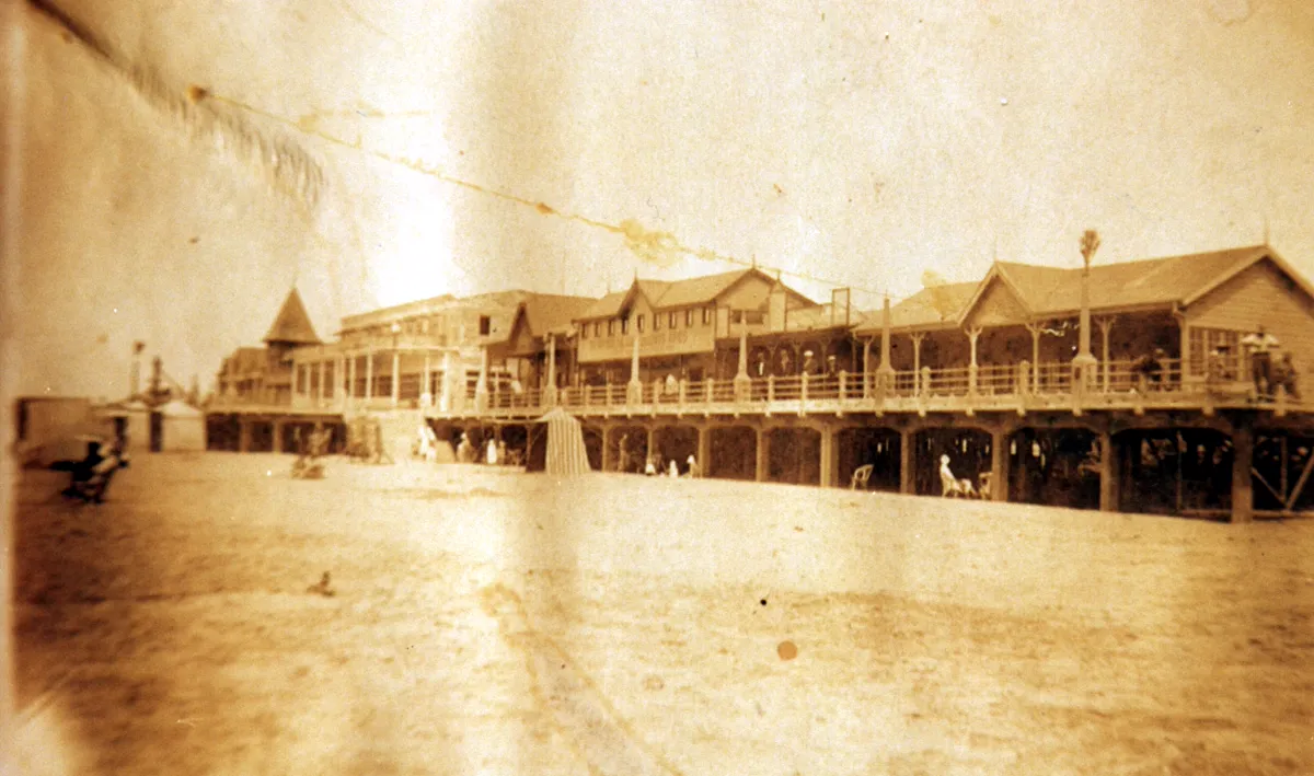 La rambla de madera vista desde la playa: pabellones largos con techos en punta sobre pilotes, una galería y una torre con techo cónico