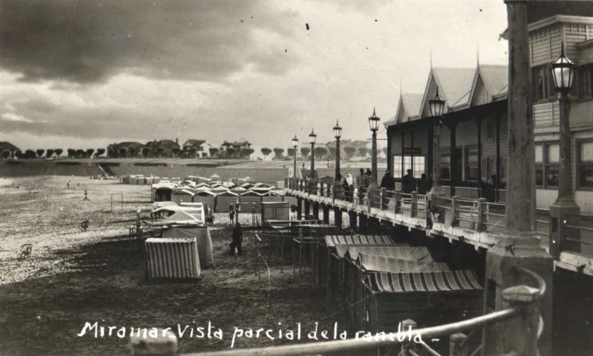 La rambla a mediados de los años 40 en un día nublado, con faroles, toldos a rayas debajo y espuma de mar volando sobre la playa